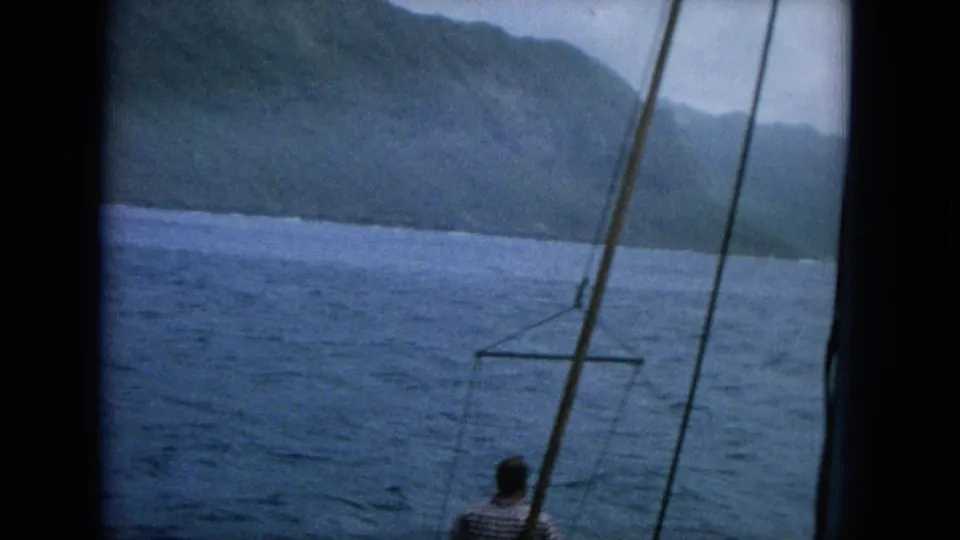 A slightly tilted 1964 Hawaii 8mm frame shows a man on a boat beneath rigging, with choppy water and mountain coastline beyond.