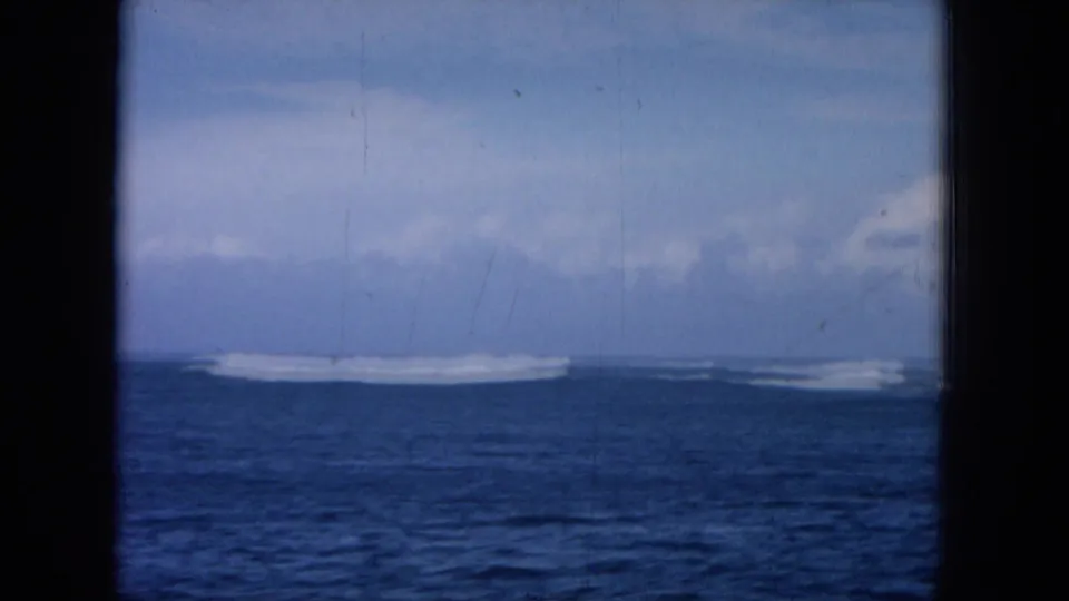 Blue offshore water and a bright surf line appear beyond a dark vertical window divider in 1964 Hawaii footage.