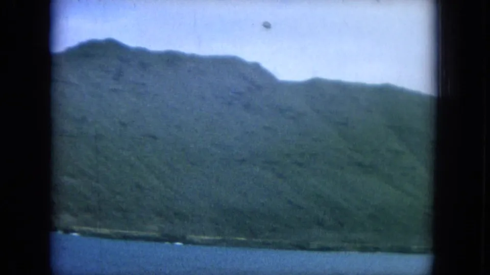 1964 Hawaii frame showing a green ridgeline along the coast, blue ocean below, and dark vertical 8mm frame borders.