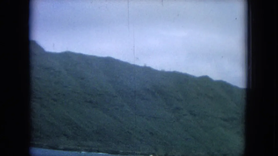 1964 Hawaii home-movie frame of a broad green mountain slope under pale sky, with ocean visible along the lower edge.