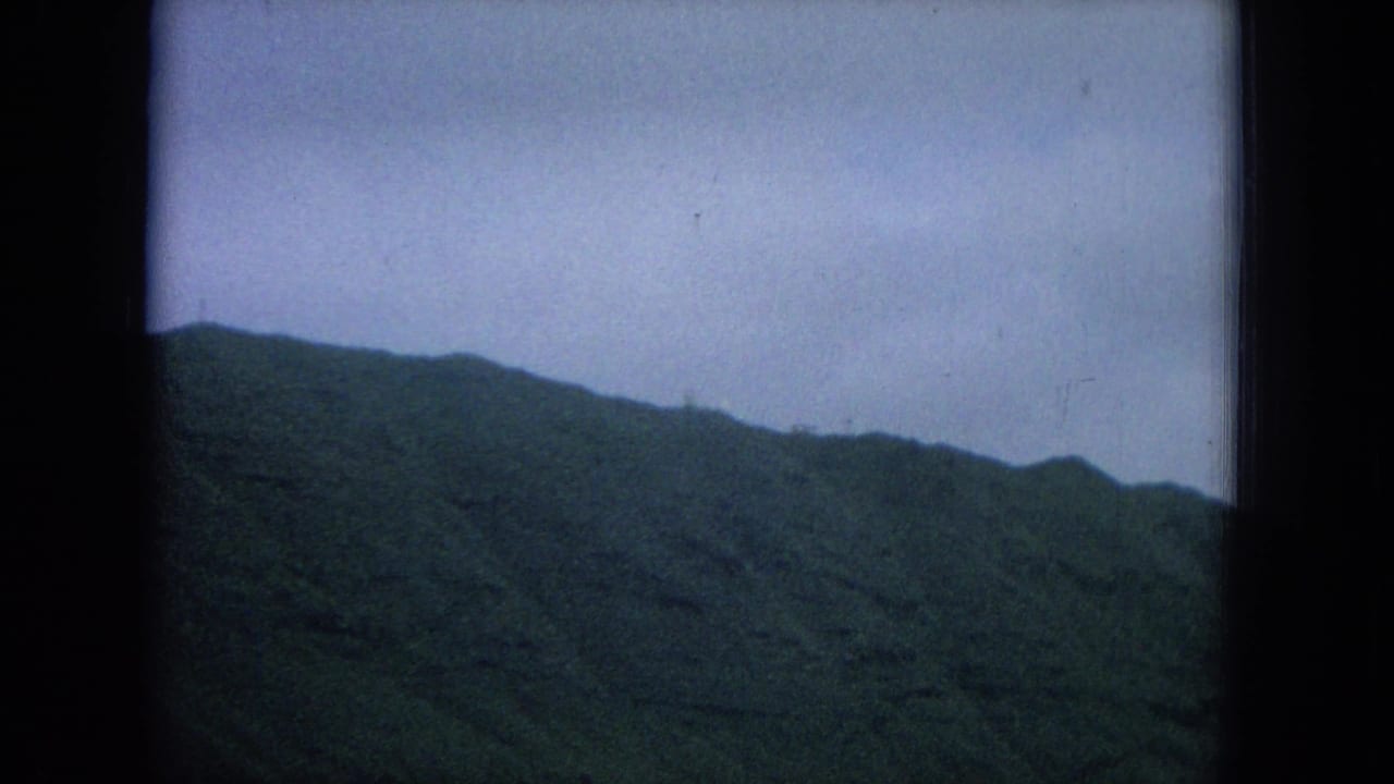 Dark green hillside beneath a pale overcast sky, framed by black 8mm film borders.