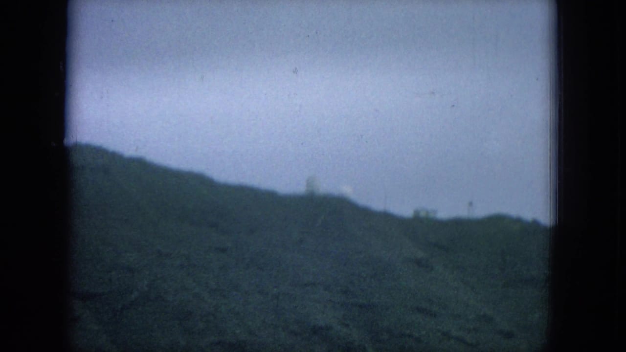Dark-edged 8mm frame of green hills beneath a pale overcast sky, with faint distant ridge shapes.