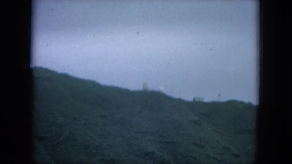1964 8mm Hawaii home-movie view of a green coastal ridge under cloudy sky, seen through dark film gate edges.