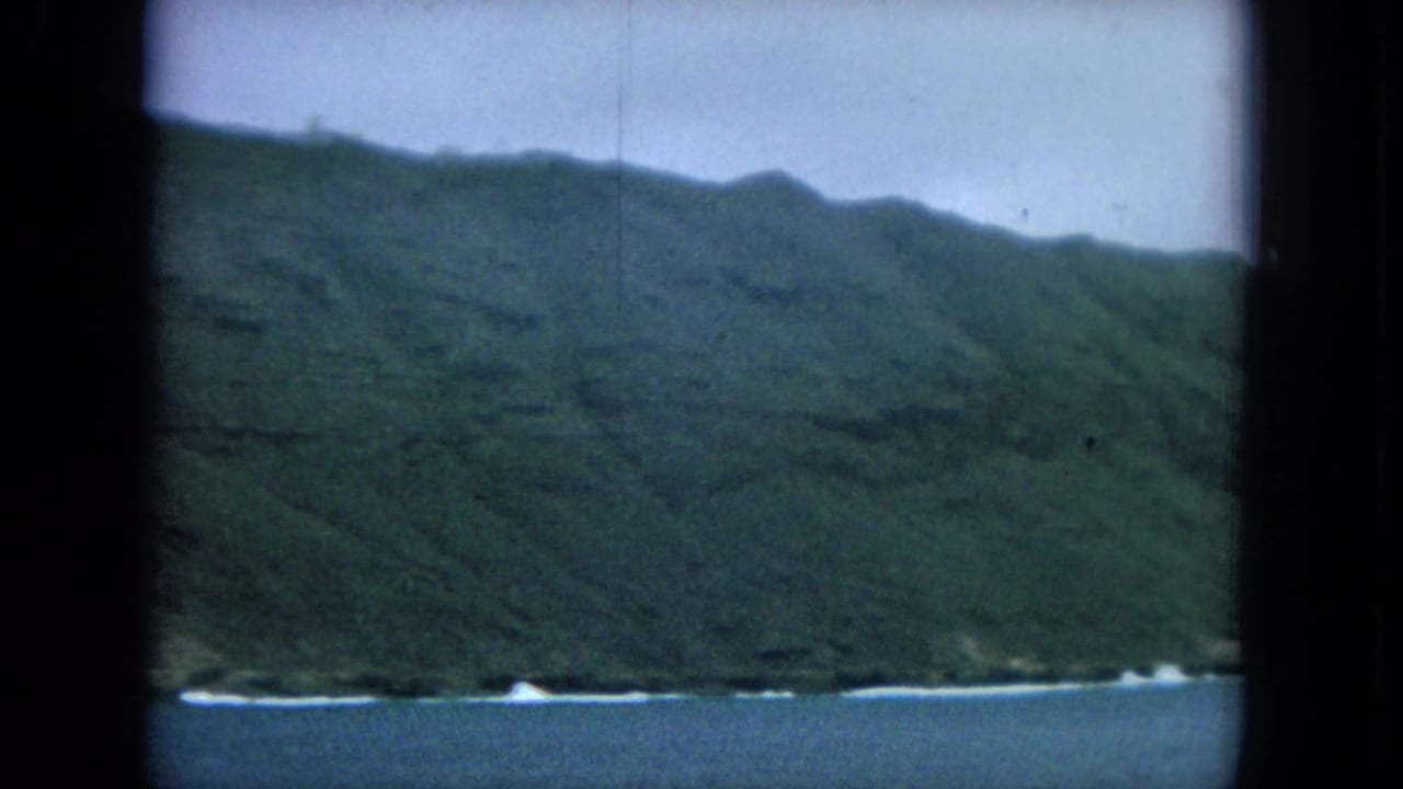 Green island hillside meets blue ocean, with white surf lines along the rocky shore.