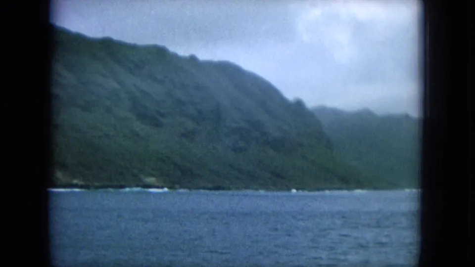 1964 8mm Hawaii boat-view coastline with green hills, blue ocean, cloudy sky, and dark film frame edges.