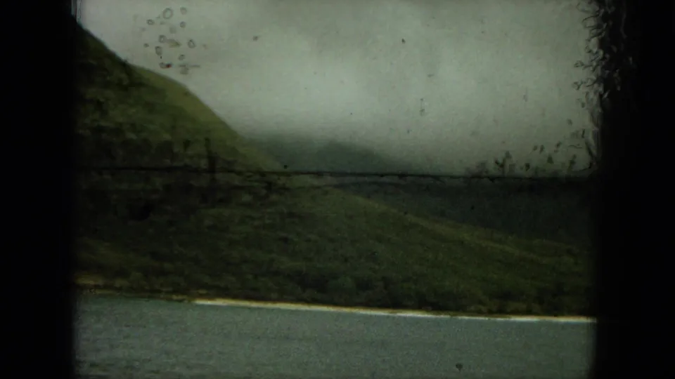 Soft 8mm frame showing a green coastal hillside, narrow pale shoreline, and muted water under hazy cloud.