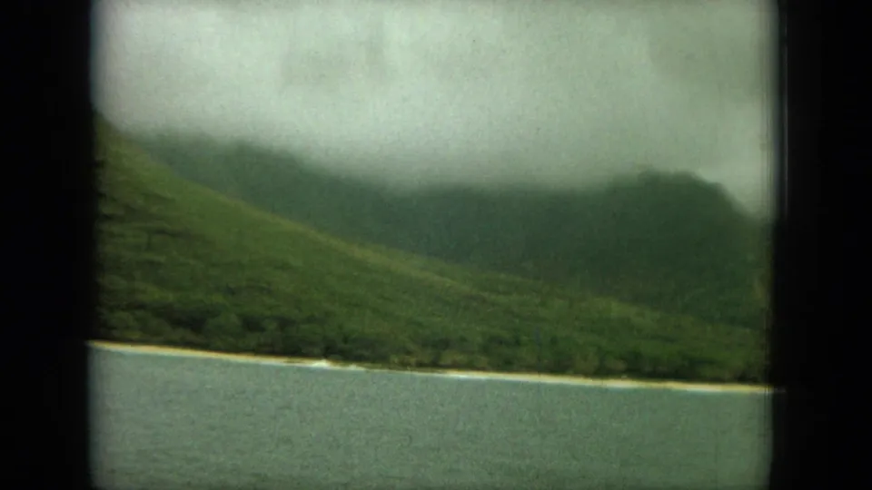 1964-style 8mm coastal view with green slopes, a narrow pale beach strip, blue-green water, and cloud cover.