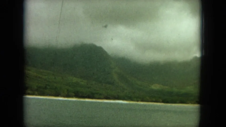 1964 Oahu home-movie frame of a clouded green ridge above a quiet coastal strip and water, with soft 8mm texture.