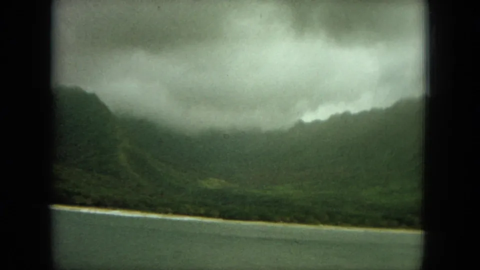 1964 Oahu 8mm frame showing a darker mountain valley, narrow coastal edge, calm water, and thick overcast clouds.