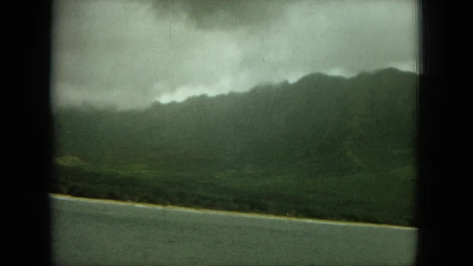 1964 Oahu home-movie frame of a misty mountain ridge above a pale shoreline and water, framed by the dark edge of the film scan.