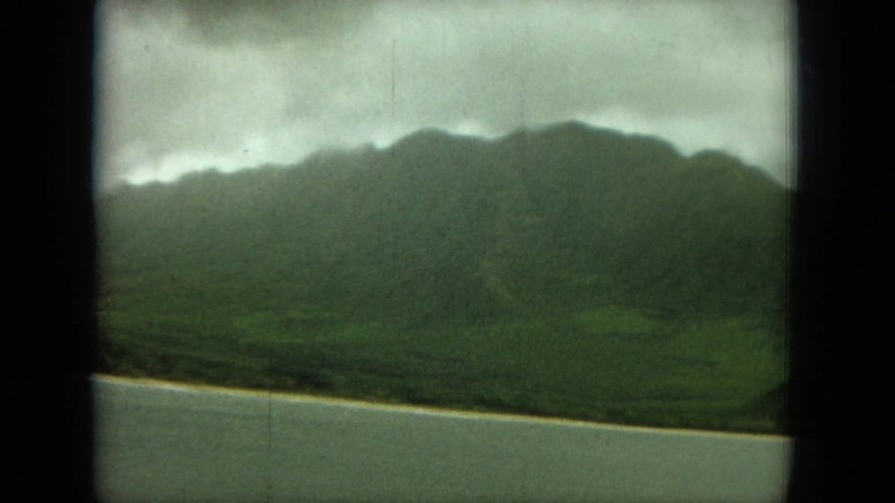 Green mountain ridge above calm coastal water under a cloudy sky.
