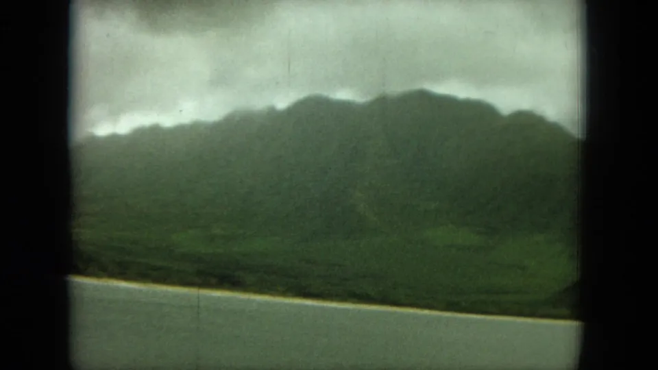 1964 Hawaii home-movie frame showing green mountains, a pale shoreline, and calm water beneath low clouds.