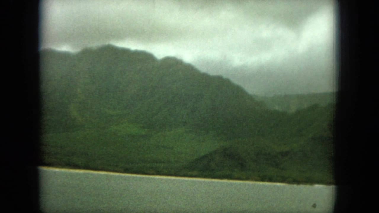 Rounded green mountains and shoreline seen across still water.