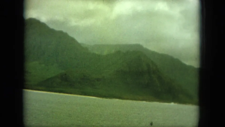 1964 Hawaii home-movie view of green hills meeting a pale coastal strip beside calm water.