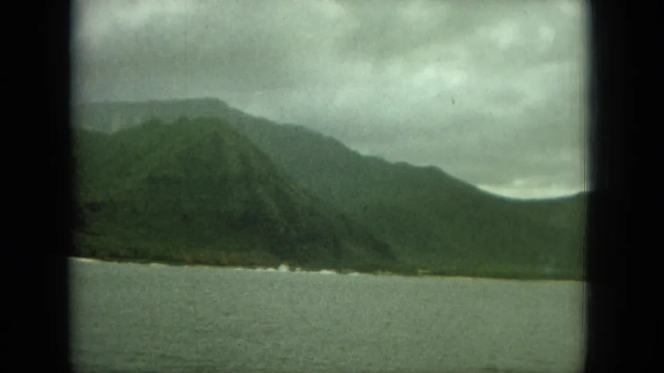 1964 Hawaii frame showing a darker mountain shoreline, calm foreground water, and heavy cloud cover.
