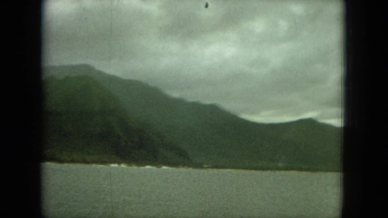 Green mountains across calm water under a cloudy sky, seen through dark window framing.