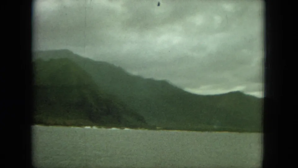 1964 8mm Oahu travel view of green mountains and calm water under overcast clouds, partially framed by a dark window edge.