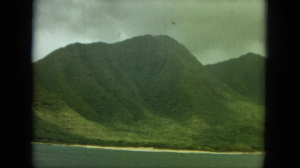 1964 Oahu home-movie frame showing a steep green ridge and narrow shoreline beside water, viewed past a dark vertical window divider.