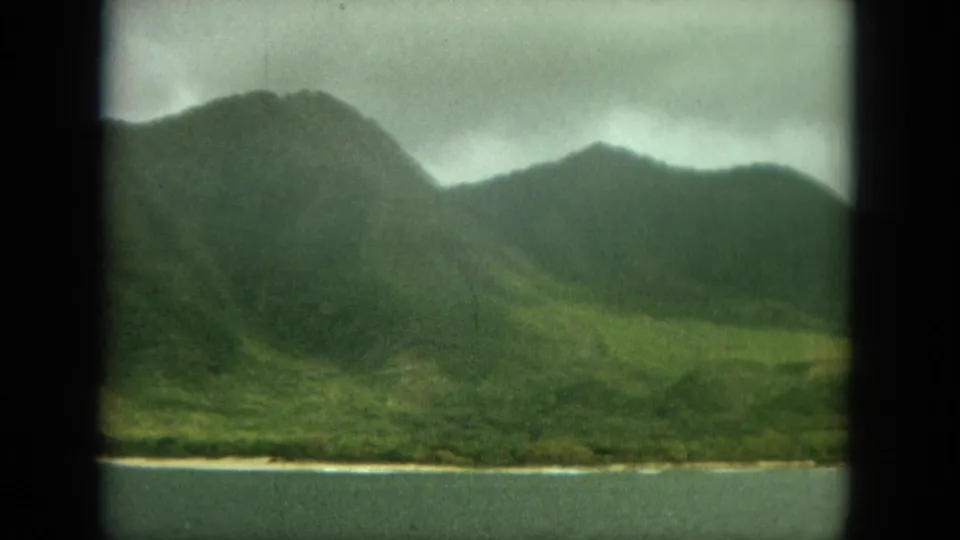 1964 8mm Oahu landscape frame with green mountain slopes, a valley, shoreline, and water seen through a windowed travel view.