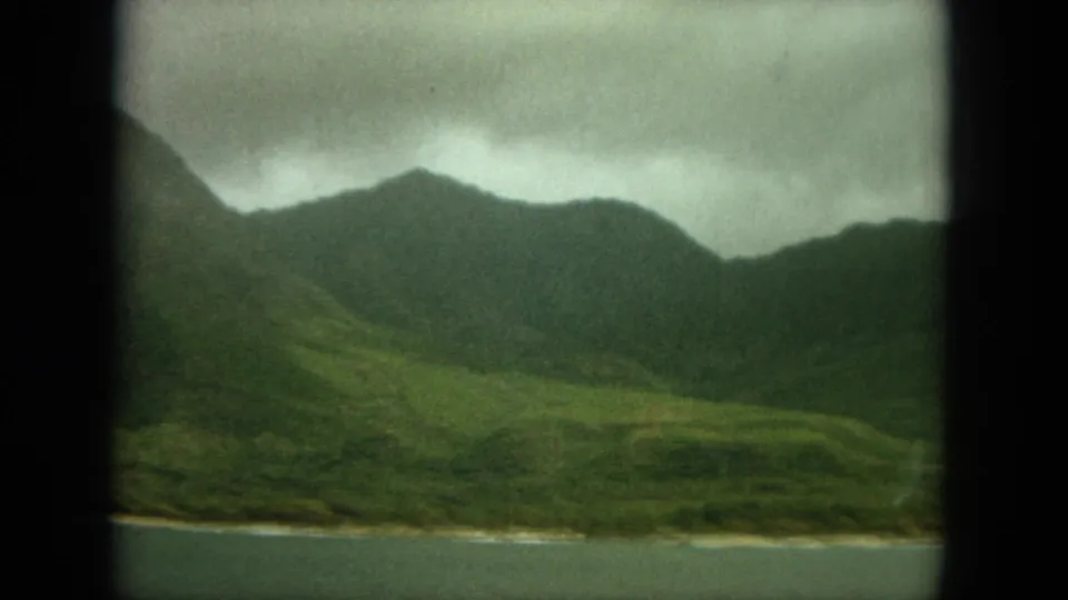 1964 Oahu travel footage frame showing a cloudy green mountain range and low shoreline across water, divided by dark window framing.