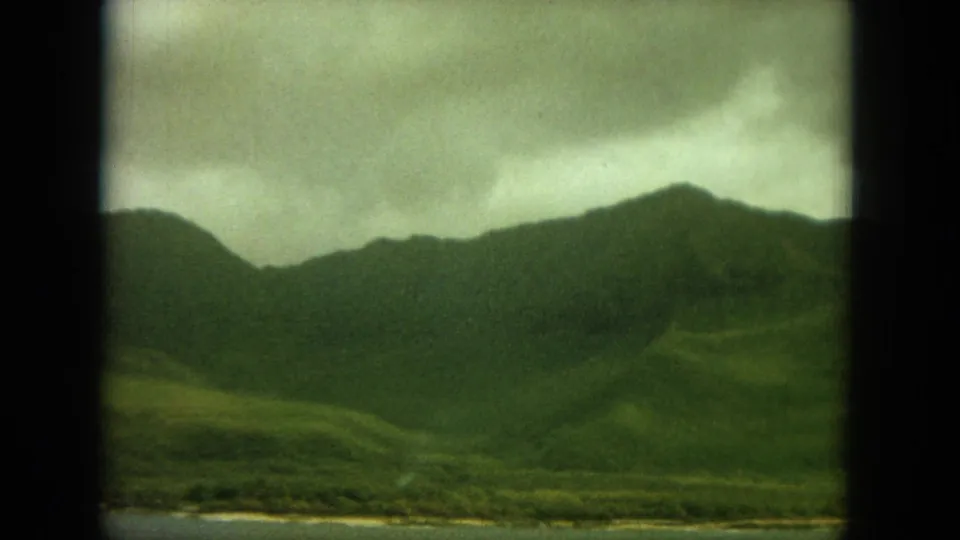 1964 Oahu landscape frame showing rounded green hills, cloudy light, a thin beach line, and calm water through an 8mm film border.