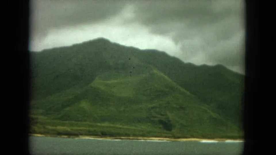 Faded 1964 8mm frame of Oahu mountain slopes, low clouds, pale shoreline, and water, with visible grain and dark frame edges.