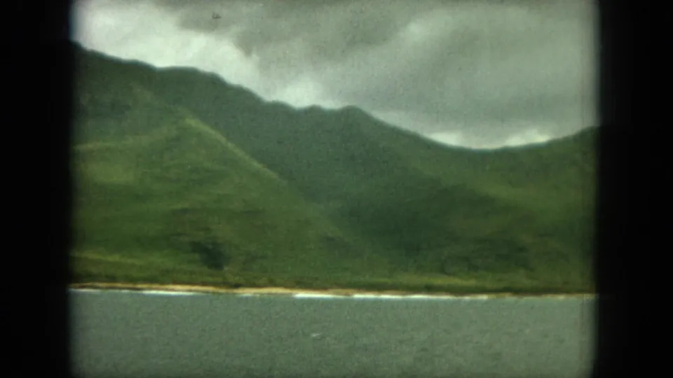 1964 Oahu coastal landscape with water in front, green ridges behind, heavy cloud cover, and dark 8mm frame borders.