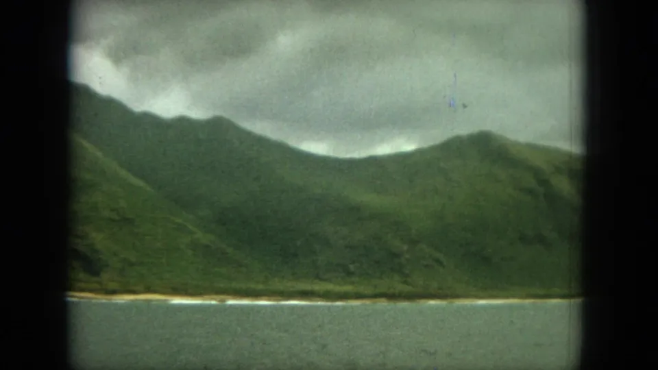 1964 8mm home-movie frame of green mountains along an Oahu coastal shoreline, seen across calm water under gray clouds.