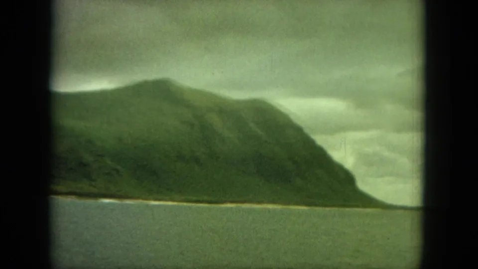 1964 8mm view of an Oahu coastal headland, with muted green slopes, a narrow shore, and cloudy sky over still water.