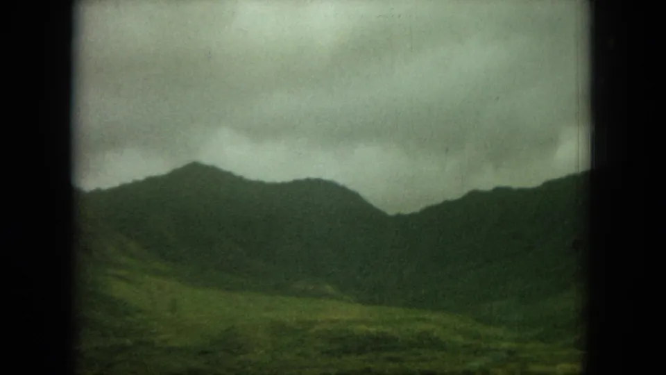 1964 8mm home-movie frame showing Oahu mountain ridges and green foreground terrain beneath an overcast sky.