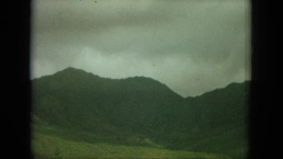 1964 8mm frame of layered Oahu mountain silhouettes, muted by haze and grain, with green terrain below cloudy skies.