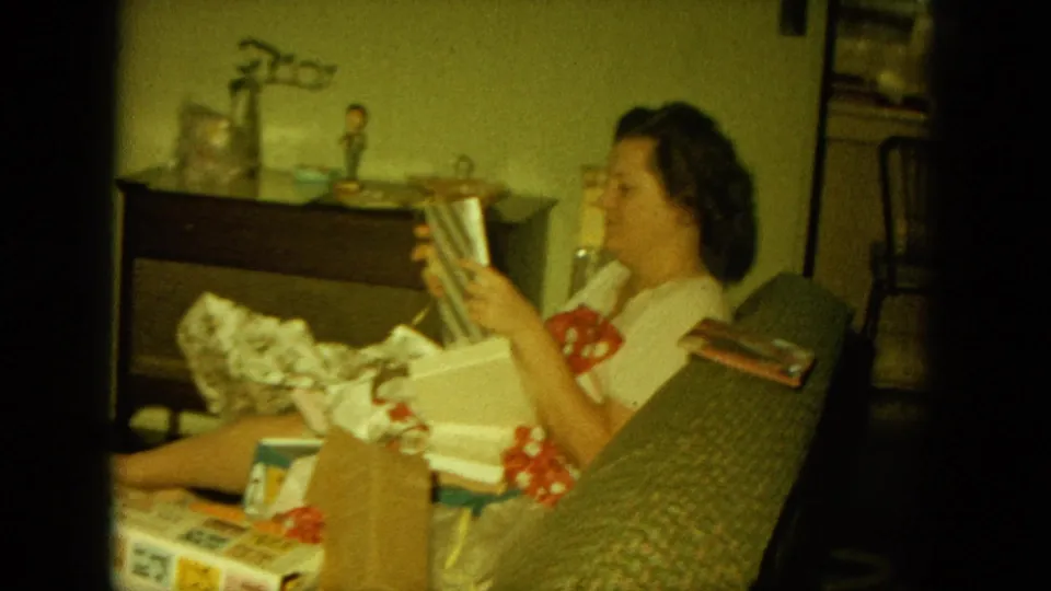 A woman in a living room holds up a striped present while seated beside holiday boxes and tissue paper.