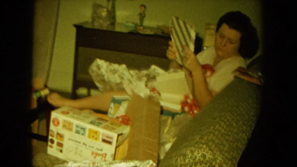 In a 1964 home-movie frame, the seated woman turns toward the camera while holding a striped Christmas gift.