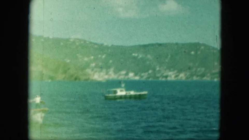 1960s 8mm frame of a small white motorboat offshore, with a green hillside and distant light buildings in the background.