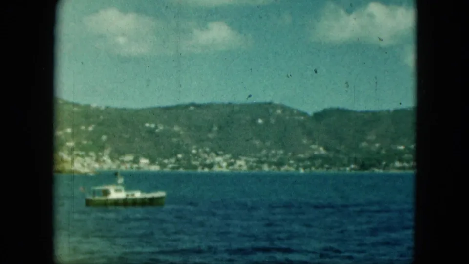Soft 8mm view of a small motorboat on open coastal water, backed by a hillside dotted with pale buildings.
