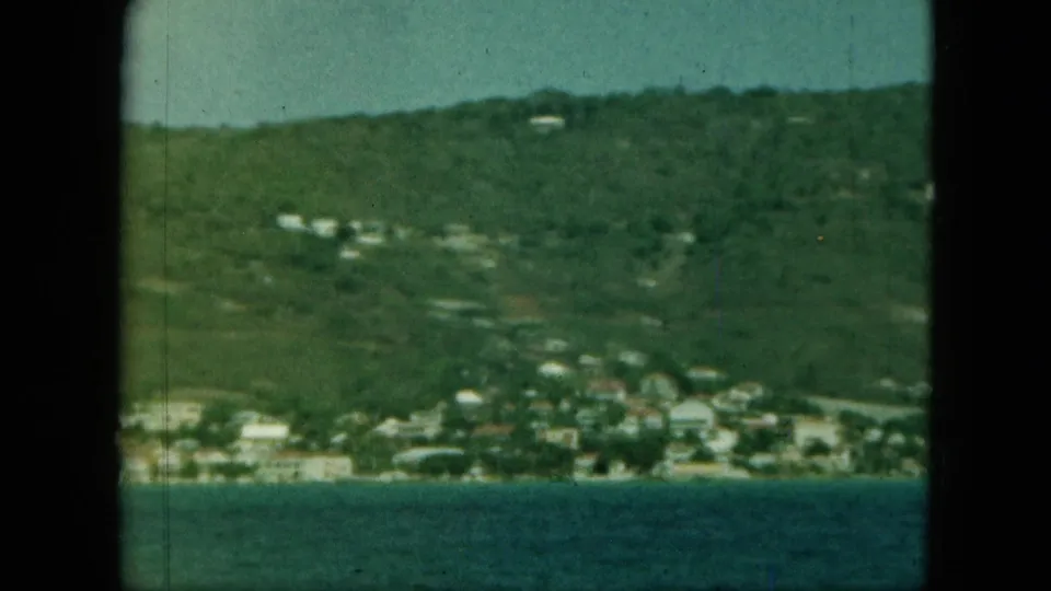 Distant coastal hillside with scattered buildings above blue water, seen through a soft, faded 8mm frame.
