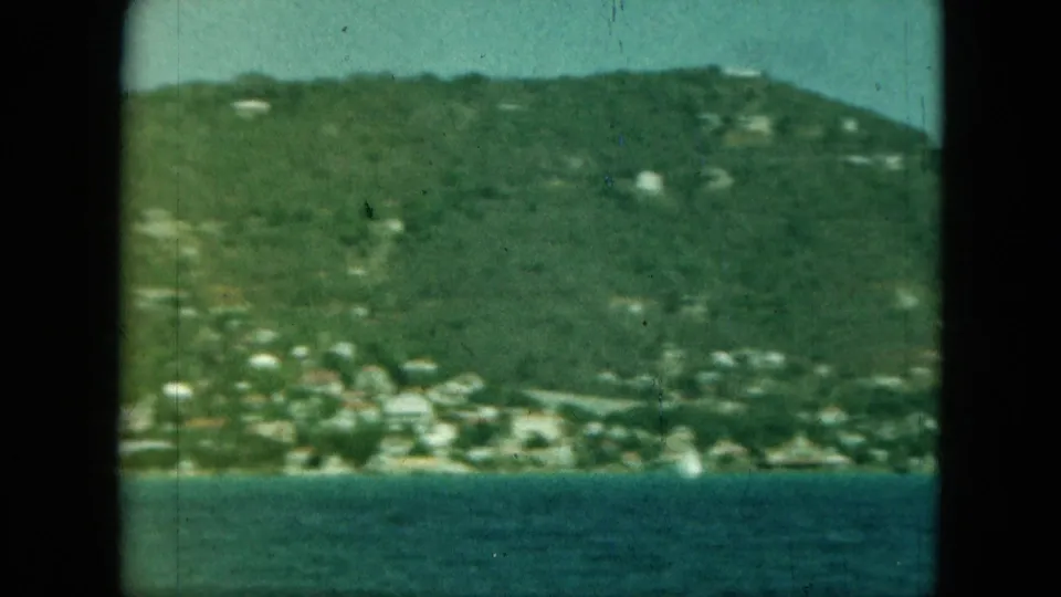 Soft 8mm frame of a green hillside and small pale buildings seen across bay water, from a likely moving visitor viewpoint.