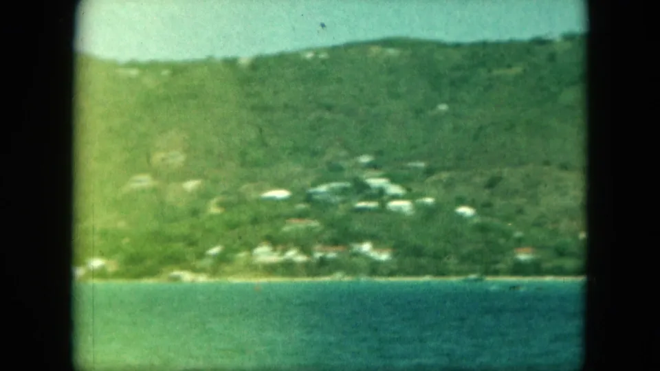 Moving 8mm view of a green island slope with scattered light buildings and bay water in the foreground.