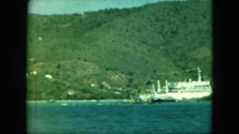 A large white ship appears near the shoreline beneath a green hillside, seen across teal water in 8mm home-movie footage.
