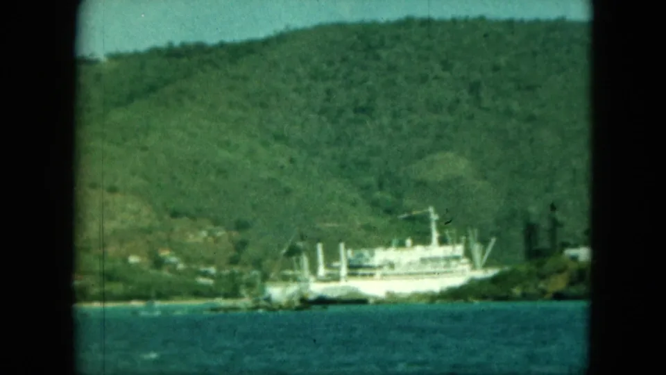 Distant white vessel moored near a tropical shoreline, with green hills rising behind and visible 8mm frame borders.