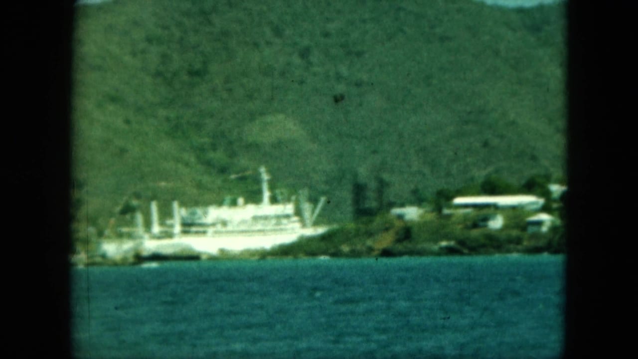 A white boat appears along the coast below green hills, with small shore structures visible in soft 8mm focus.