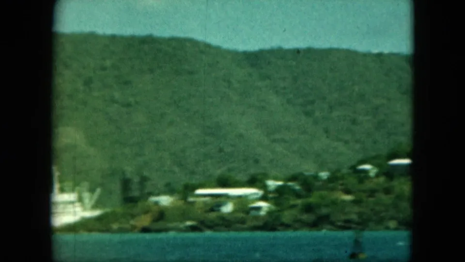 The panning view catches the edge of the vessel and shoreline buildings, with water below and green hillside filling the background.