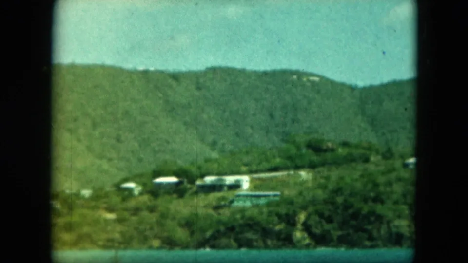 Small coastal buildings and vegetation line the shore, with blue water in front and a green hillside under bright sky.