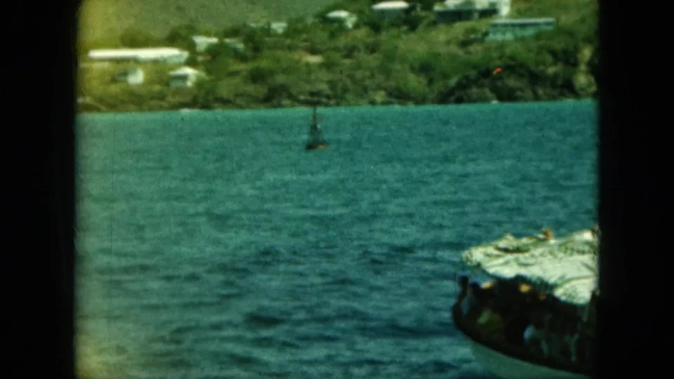 1964 8mm-style view from a moving boat toward a green shoreline, with a small passenger boat crossing the foreground.