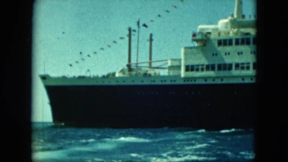 Large ocean liner seen close from the water, with white superstructure, dark hull, funnels, and signal flags overhead.