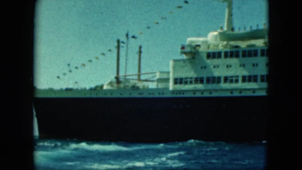 Handheld 8mm frame showing the side of a large ocean liner, with rows of windows, signal flags, and wake at the hull.