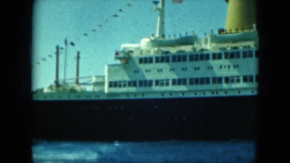 Close 1960s home-movie view of a passenger ship’s white decks, yellow funnel, dark hull, and string of signal flags.