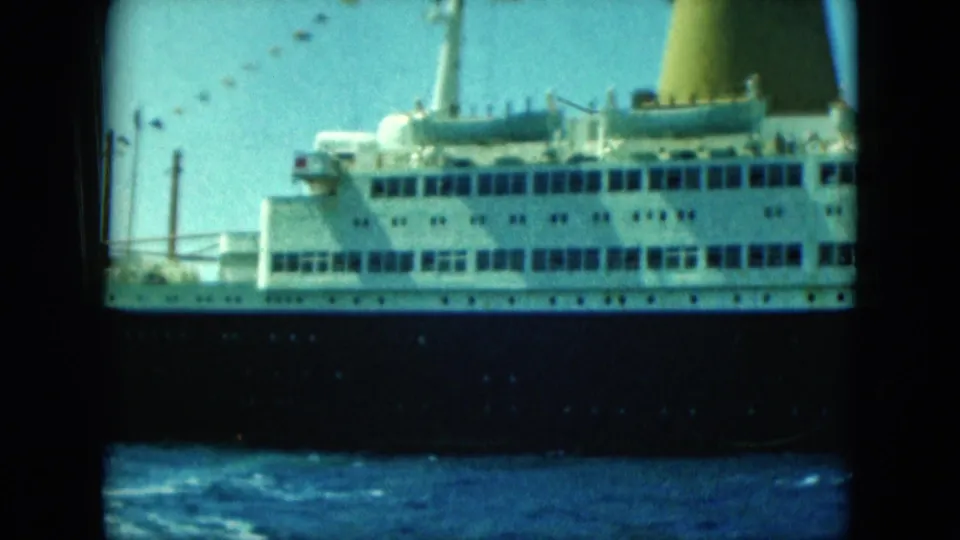 A large cruise ship with a white upper deck and dark hull moves across the sea in archival 8mm footage dated 1964 by metadata.