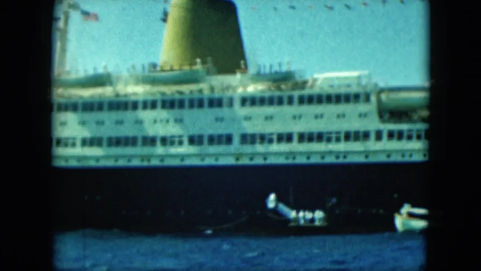 A large cruise ship sits beyond two small boats, including a tender carrying people, in soft blue-green 8mm footage.