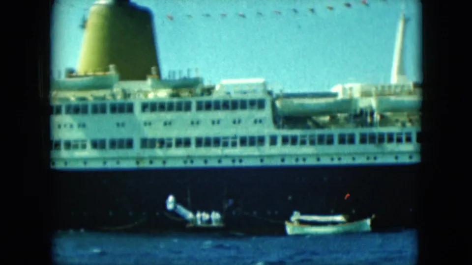 The ship remains broadside in frame, with small boats on the water below and a pale vertical structure visible near the right edge.